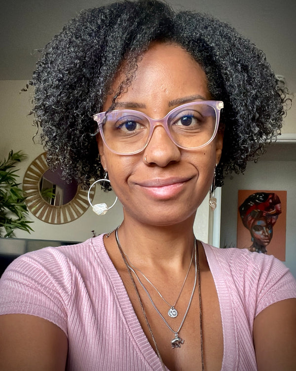 African American woman with glasses, curly hair a light pink top, citrine silver wire wrapped earrings and 2 silver chains looking at the camera and smiling.